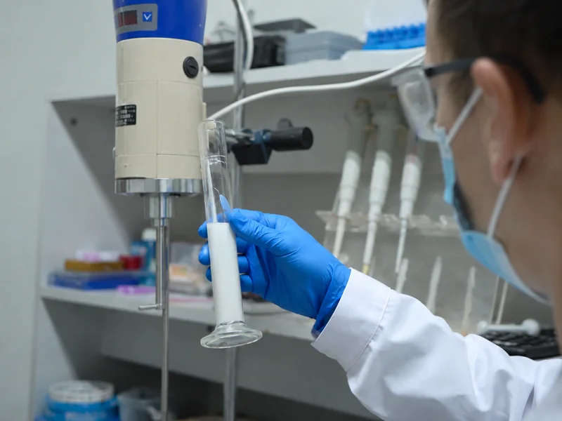 Acercamiento creado con IA: una mano con guante sostiene una muestra de recubrimiento junto a un mezclador de laboratorio.